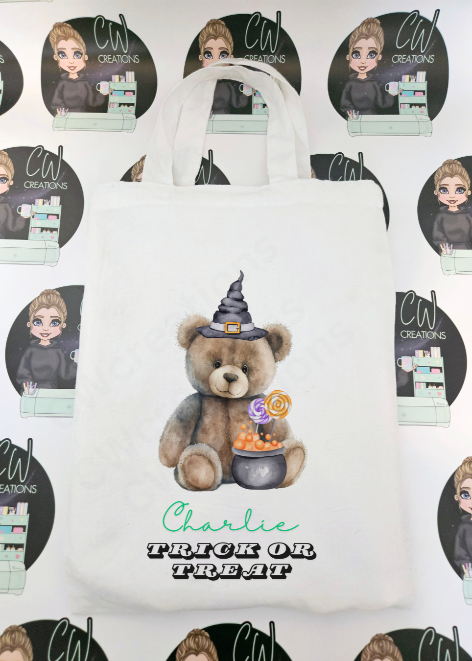 A white bag with a teddy wearing a witches hat sitting in front of a cauldron.