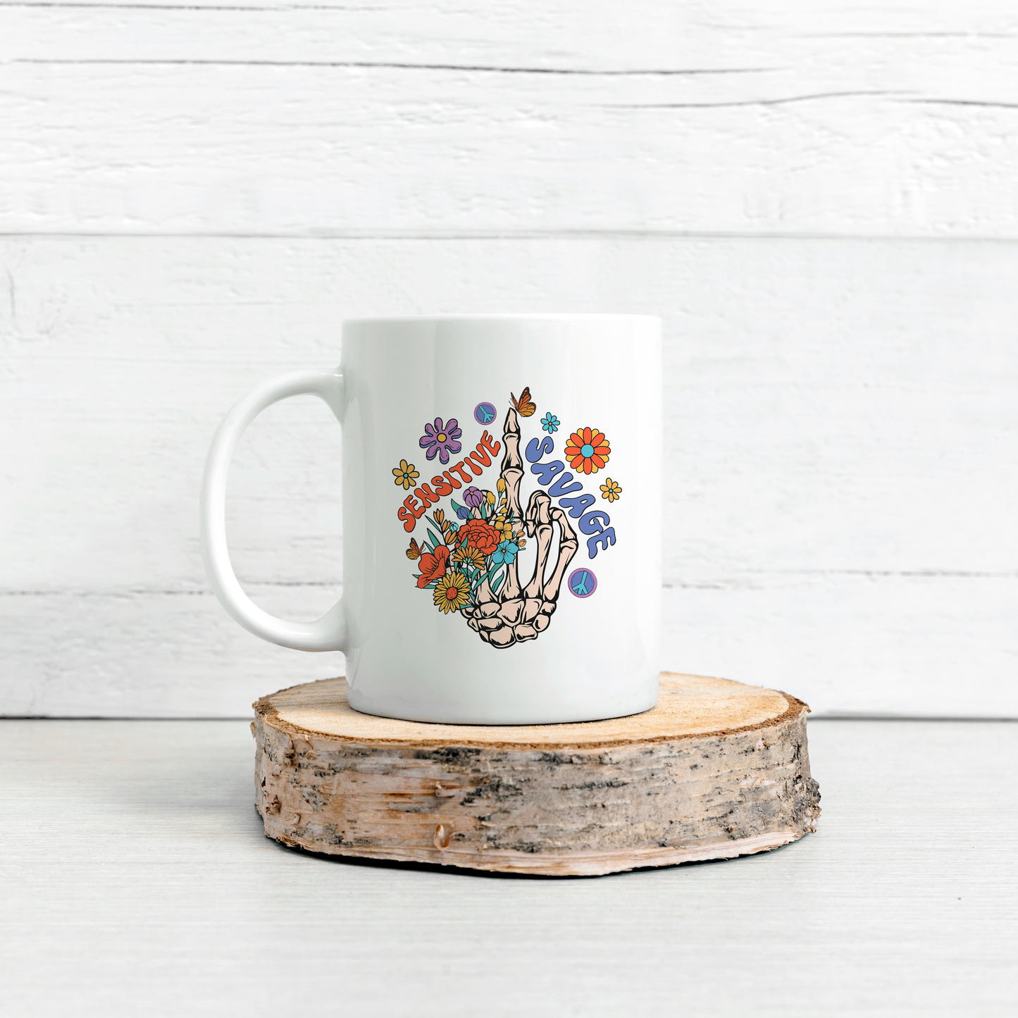 White mug with the words Sensitive Savages with a graphic showing a skeleton hand showing middle finger with bright coloured flowers. The white mug is displayed on sliced log with a white brick walled background.