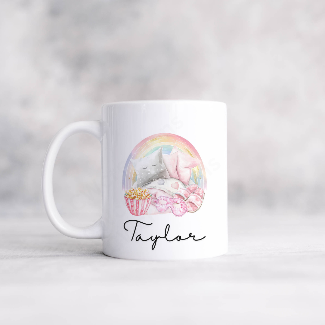 A white mug with a pink sleep over style graphic with a rainbow, pillows, popcorn, slippers personalised with a name.